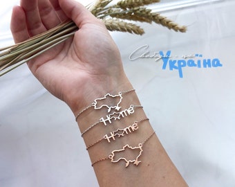 Könnte beinhalten: Ein Set aus drei Armbändern mit einer Umrandung der Ukraine und dem Wort "Home" auf jedem. Die Armbänder befinden sich am Handgelenk einer Person. Der Hintergrund ist eine weiße Oberfläche mit Weizenhalmen.
