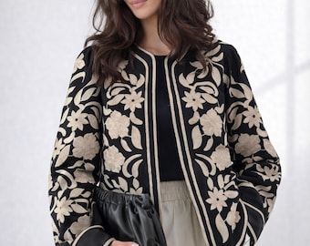 May include: Black jacket with cream-colored floral embroidery. The jacket has a cream-colored trim and cuffs. A black clutch is held in the hand. The jacket is worn over a black top and a light-colored skirt.