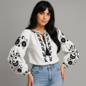 May include: A white long-sleeved blouse featuring black floral and bird embroidery on the sleeves and front placket. The garment has a round neckline with a central black tassel tie closure and voluminous sleeves with fitted cuffs.