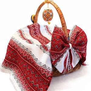 Puede incluir: Una cesta de mimbre tejida con asa, forrada con un paño blanco con un patrón bordado en rojo y negro. Un lazo decorativo hecho con la misma tela está atado a la cesta. Un huevo decorado cuelga del asa.