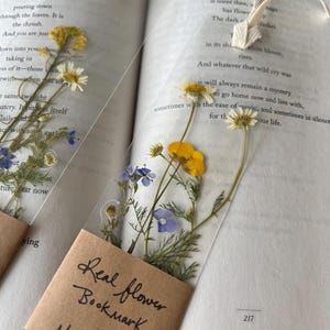 May include: Two clear bookmarks with pressed flowers, including yellow, white, and blue blooms. Each bookmark has a small brown paper tag that reads "Real flower Bookmark Made in PA". The bookmarks are placed in an open book.