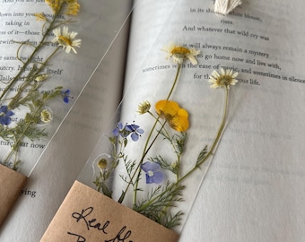 Può includere: Due segnalibri trasparenti con fiori pressati gialli, bianchi e blu. Ogni segnalibro ha un piccolo cartellino di carta marrone con la scritta "Real flower Bookmark Made in PA". I segnalibri sono in un libro aperto.