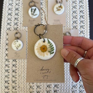 May include: A white ceramic keychain with a pressed daisy and green leaves. The keychain is attached to a brown card with the text "daisy a symbol of joy" and the website "wildflowerandjanco.etsy.com".