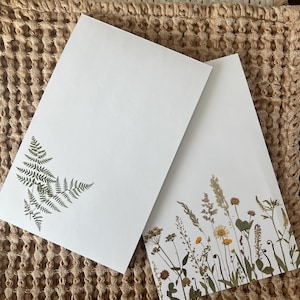 May include: Two white stationery cards with botanical designs. One card features a fern print in the corner, while the other showcases a variety of dried flowers and grasses along the bottom edge. The cards are set against a textured, neutral-toned background.