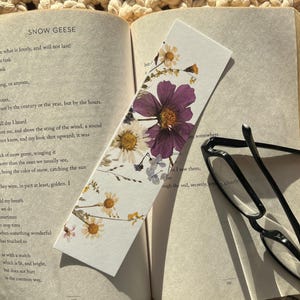 Könnte beinhalten: Ein Lesezeichen mit gepressten Blumen in Weiß, Gelb und Lila. Das Lesezeichen liegt auf einem Buch mit dem sichtbaren Text "SNOW GEESE".