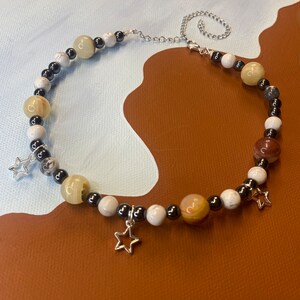 Puede incluir: Un collar de cuentas blancas, marrones y negras con abalorios de estrellas plateadas. El collar está hecho con varias piedras y cuentas, incluyendo howlite blanca, ágata marrón y hematites negra.