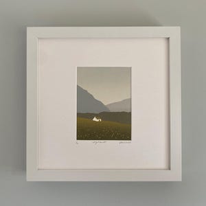 May include: A framed art print featuring a minimalist landscape scene. The artwork depicts a small white house nestled in a green field, with mountains in the background. The print is in a white frame.