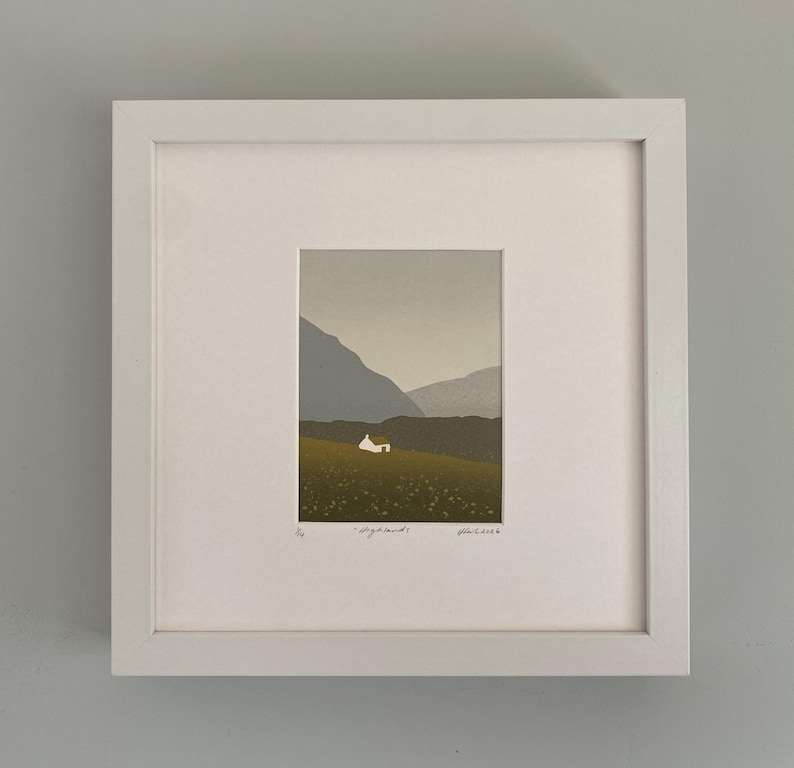 May include: A framed art print featuring a minimalist landscape scene. The artwork depicts a small white house nestled in a green field, with mountains in the background. The print is in a white frame.