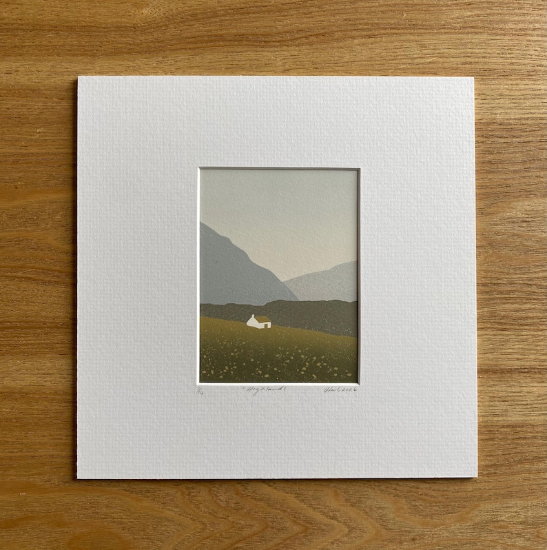 May include: A framed print of a landscape scene featuring a small white house on a green hillside, with gray mountains in the background. The print is set against a white mat and is on a wooden surface.