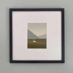 May include: Framed art print featuring a minimalist landscape scene. A small white house sits on a green hillside, with mountains in the background under a muted sky. The print is set within a white mat and a black frame.