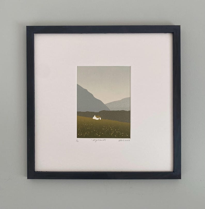 May include: Framed art print featuring a minimalist landscape scene. A small white house sits on a green hillside, with mountains in the background under a muted sky. The print is set within a white mat and a black frame.