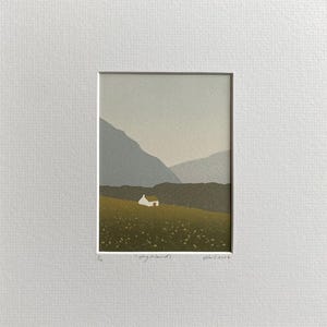 May include: A framed print of a landscape featuring a small white house on a green hillside, with mountains in the background. The sky is a pale gray, and the print is mounted on a white mat.