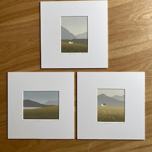 May include: Three framed landscape prints. Each print features a small white house in a field of yellow flowers, with mountains in the background. The prints are matted in white and displayed on a wooden surface.