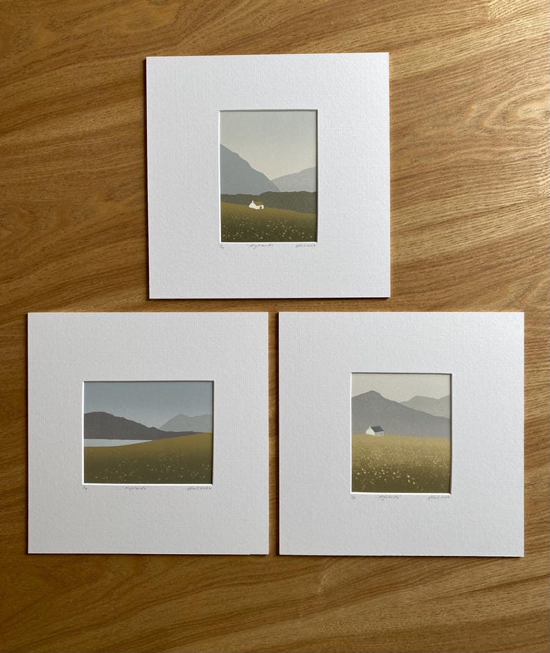 May include: Three framed landscape prints. Each print features a small white house in a field of yellow flowers, with mountains in the background. The prints are matted in white and displayed on a wooden surface.