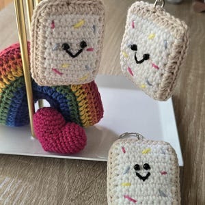 May include: Crocheted keychains shaped like smiling pastries with colorful sprinkles. The keychains have a tan border and a white center. A crocheted rainbow and a pink heart are also visible.