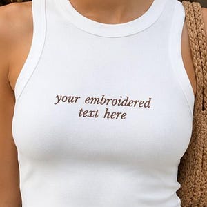 Op de afbeelding: Een wit mouwloos hemdje met de bruin geborduurde tekst "your embroidered text here" op de borst, gecombineerd met een donkerbruine, losvallende broek met een elastische tailleband met trekkoord en een gevlochten bruine schoudertas.