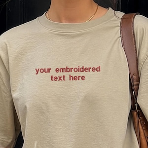 May include: A beige short-sleeved t-shirt featuring the embroidered text "your embroidered text here" in a dark red font across the chest. The shirt has a crew neckline and a relaxed fit, paired with blue denim jeans and a brown shoulder bag.