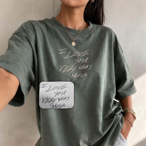 Può includere: Una maglietta verde oliva con la scritta a mano "I LOVE YOU VERY VERY MUCH" in bianco. La maglietta ha una vestibilità ampia e maniche corte. È visibile anche un piccolo biglietto scritto a mano con lo stesso testo.