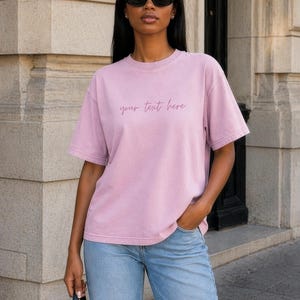Peut inclure: Un t-shirt à manches courtes et col rond de couleur mauve atténuée. Le vêtement présente le texte "your text here" imprimé dans une fine police cursive au centre de la poitrine. Il possède une coupe décontractée avec un ourlet standard.