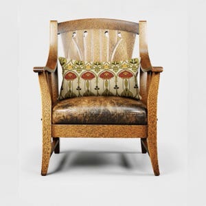 May include: A wooden armchair with a dark brown leather seat and a slatted backrest. A rectangular throw pillow rests on the seat, featuring a repeating pattern of red, green, and yellow floral motifs on a light cream-colored background.