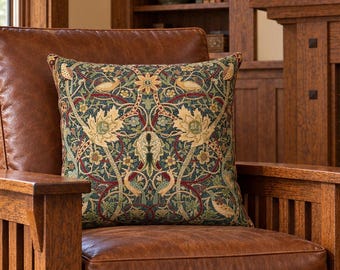 May include: A brown leather armchair with a wooden frame featuring vertical slats. A square throw pillow rests on the seat, displaying a detailed botanical pattern with birds, leaves, and flowers in shades of green, gold, and deep red.