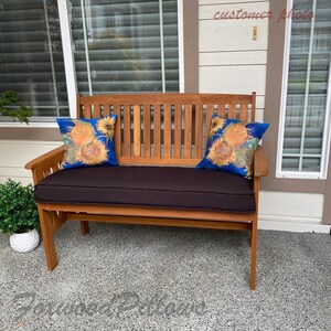 Pu&ograve; includere: Una panca di legno con un cuscino marrone e due cuscini decorativi con motivo di girasole. La panca &egrave; su un patio con pavimento in cemento.