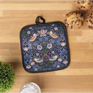 May include: A square, navy blue pot holder with a black border and a hanging loop. The pot holder features a floral and bird design in shades of blue, pink, and green. The design includes birds, flowers, and berries. The pot holder is on a wooden surface.