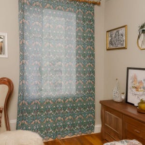 Pu&ograve; includere: Una tenda trasparente con un motivo ripetuto di uccelli ed elementi floreali nei toni del verde acqua, pesca e bianco. La tenda &egrave; appesa davanti a una finestra, aggiungendo un tocco decorativo alla stanza.
