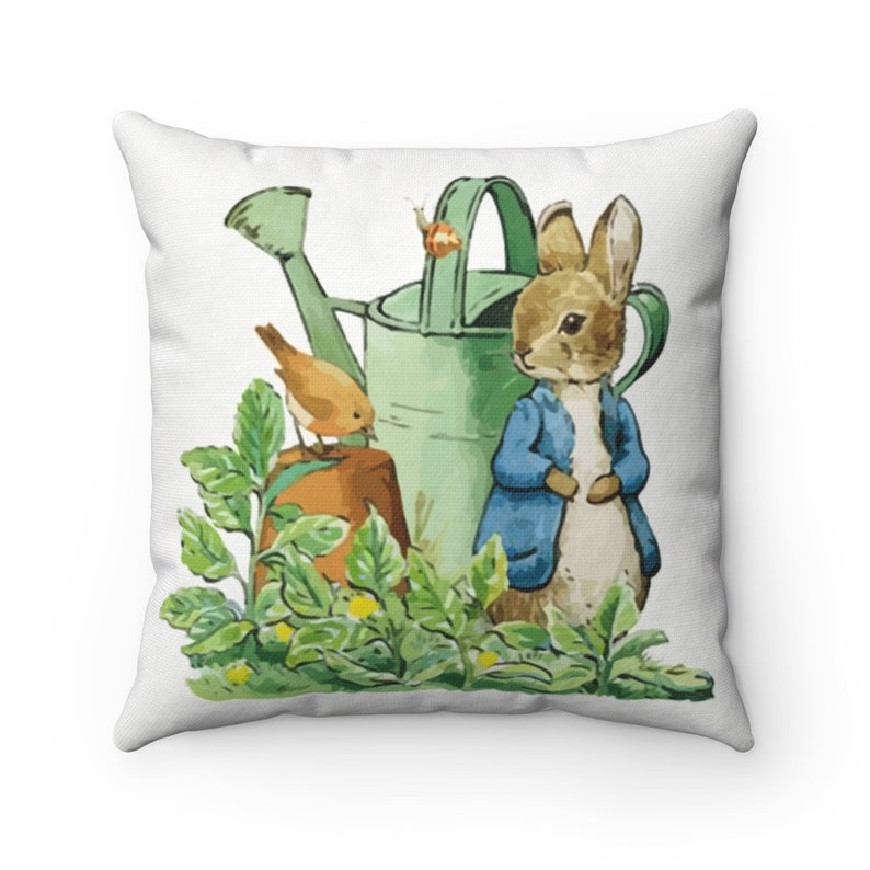 May include: A white throw pillow with a green watering can, a brown rabbit wearing a blue jacket, and a small orange bird perched on a brown pot. The background is a white fabric with green leaves.