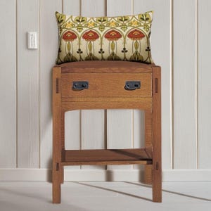 May include: A rectangular throw pillow featuring a repeating floral pattern in shades of red, yellow, and green on a cream background, resting on a wooden nightstand with one drawer, two dark metal handles, and an open lower shelf.