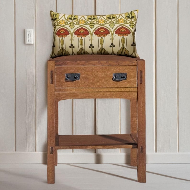 May include: A rectangular throw pillow featuring a repeating floral pattern in shades of red, yellow, and green on a cream background, resting on a wooden nightstand with one drawer, two dark metal handles, and an open lower shelf.