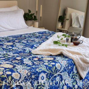 May include: A blue and white floral patterned bedspread with a white towel on top. The bedspread has a design of flowers and leaves. The towel is folded in half and has a green plant on top. The text "FOXWOOD PILLOWS" is visible at the bottom of the image.