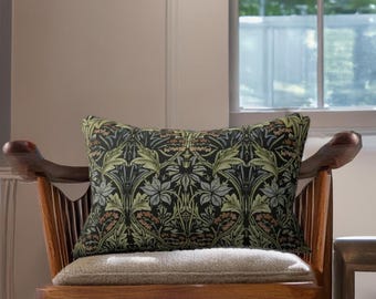 May include: A rectangular cushion with a dark background and a repeating floral pattern in green, grey, and brown. The cushion sits on a wooden chair with a light-coloured cushion. The chair is in front of a window.