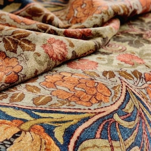 May include: Close-up of a fabric with a floral pattern in shades of orange, brown, blue, and beige. The design features stylized flowers and leaves, creating a textured, layered effect. The fabric appears soft and draped.