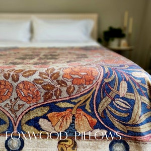 May include: A close-up of a patterned throw blanket with a floral design in shades of blue, orange, and brown. The blanket is on a bed with white pillows and a wooden headboard. The text "FOXWOOD PILLOWS" is visible in the bottom of the image.