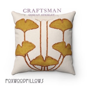 Puede incluir: Una almohada blanca con un diseño estilizado de hojas de ginkgo en marrón y amarillo. La almohada presenta el texto "CRAFTSMAN GUSTAV STICKLEY" en morado.