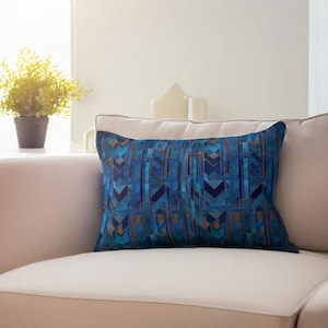 Könnte beinhalten: Ein rechteckiges Zierkissen mit einem geometrischen Muster in Blau- und Bronzetönen. Das Kissen liegt auf einem hellen Sofa. Das Design zeigt vertikale Linien und eckige Formen.