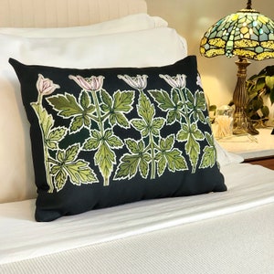 May include: A black pillow with a floral design featuring white and pink flowers and green leaves. The pillow is on a white bed with a white headboard and a lamp with a stained glass shade.