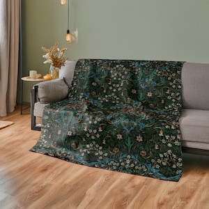 May include: A dark green throw blanket with a floral pattern, draped over a gray sofa. The blanket features a repeating design of white, blue, and red flowers and foliage. The sofa is in a room with wooden floors and a light green wall.