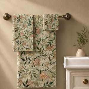 May include: A set of three towels hanging on a bronze-toned metal bar, featuring a repeating botanical pattern with light green leaves, dark green stems, and clusters of small, pale orange flowers against a light cream-colored background.