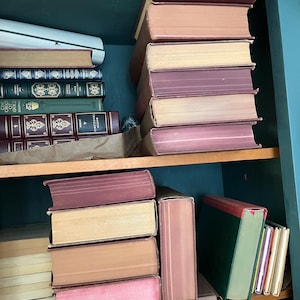 May include: A collection of antique books with various colored spines and aged pages, stacked on wooden shelves. The books have shades of pink, tan, and burgundy. The shelves are painted a teal color.