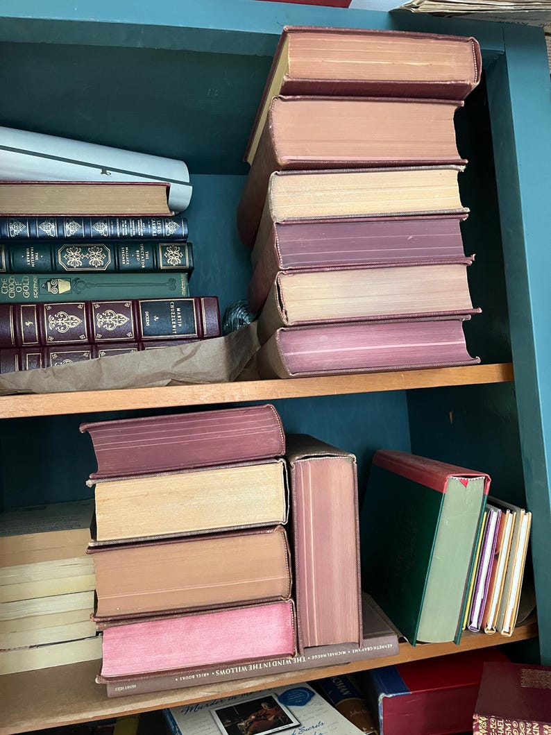 May include: A collection of antique books with various colored spines and aged pages, stacked on wooden shelves. The books have shades of pink, tan, and burgundy. The shelves are painted a teal color.
