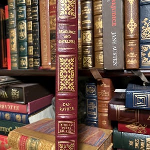 May include: A burgundy hardcover book with gold lettering and a gold design on the cover. The title of the book is "Deadlines and Datelines" by Dan Rather. The book is a signed first edition.