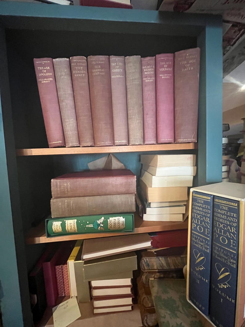 May include: A teal bookshelf filled with vintage books. The top shelf displays a row of maroon-colored books with gold lettering. The middle shelf holds stacked books in various colors, including a green book. The bottom shelf is partially visible, with more books.
