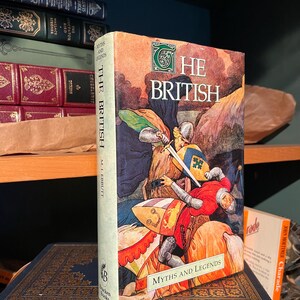 May include: A hardcover book titled "The British Myths and Legends" with a colorful illustration of knights in battle. The spine reads "The British" and "M. LEBBUTT". The book is set against a backdrop of other books.