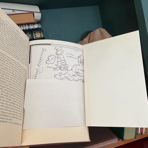 May include: An open book with a map illustration of Europe, including the British Isles, and text in English. The book is on a shelf with other books, with a green and brown background.