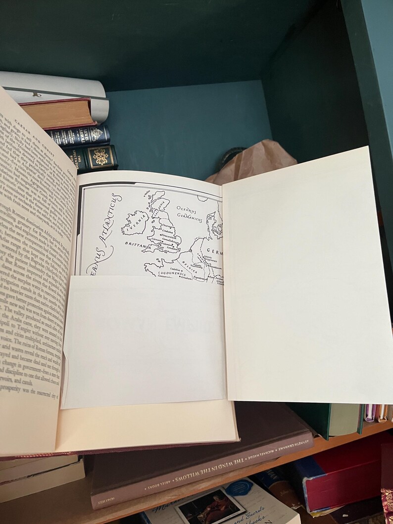 May include: An open book with a map illustration of Europe, including the British Isles, and text in English. The book is on a shelf with other books, with a green and brown background.