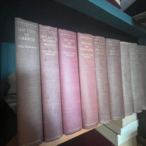 May include: A shelf displays a collection of vintage books with titles like "The Life of Greece" and "The Age of Reason Begins." The books have textured covers in shades of red and tan, with gold lettering. The books are arranged on a wooden shelf.