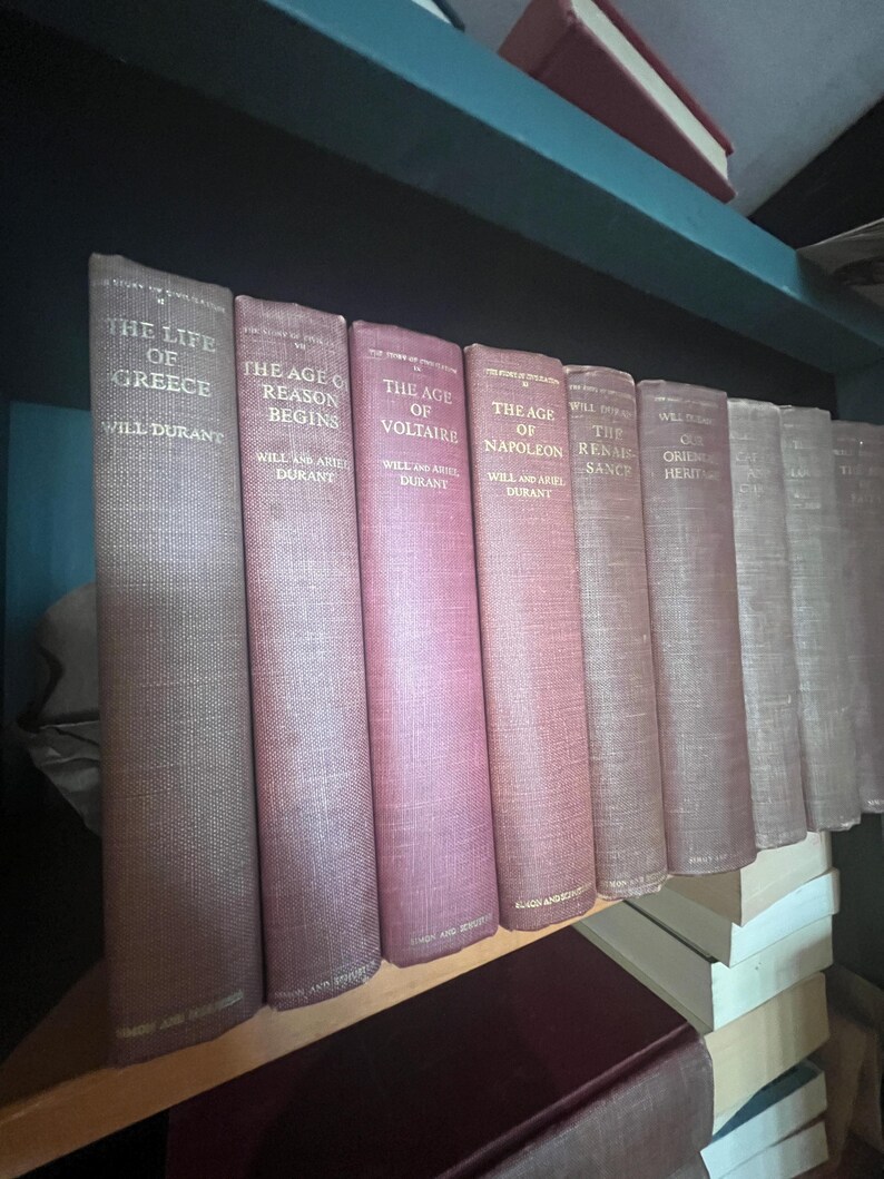 May include: A shelf displays a collection of vintage books with titles like "The Life of Greece" and "The Age of Reason Begins." The books have textured covers in shades of red and tan, with gold lettering. The books are arranged on a wooden shelf.