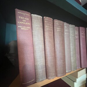 May include: A collection of antique books with maroon and tan covers, arranged on a wooden shelf. The spines display titles like "The Age of Napoleon" and "Caesar and Christ." The books are part of a historical collection.